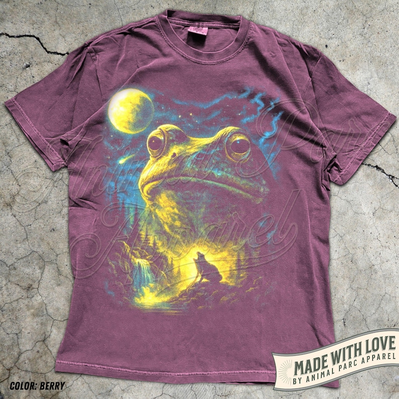 May include: A berry-colored short-sleeve t-shirt featuring a graphic print of a large frog looking up at a yellow moon in a starry night sky. Below the frog, a small wolf silhouette stands on a rocky landscape near a waterfall. The shirt has a crew neckline.