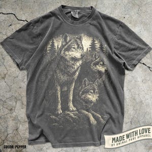 May include: Pepper-coloured t-shirt with a detailed illustration of three wolves in a forest setting. The design includes a light-coloured background with trees and stars. The text "MADE WITH LOVE BY ANIMAL PARC APPAREL" is printed on a banner.