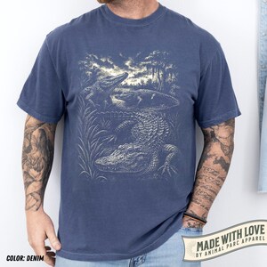 May include: Denim blue t-shirt featuring a detailed illustration of alligators in a swamp setting. The graphic is in a vintage style, with the text "MADE WITH LOVE BY ANIMAL PARC APPAREL" on a banner at the bottom right.