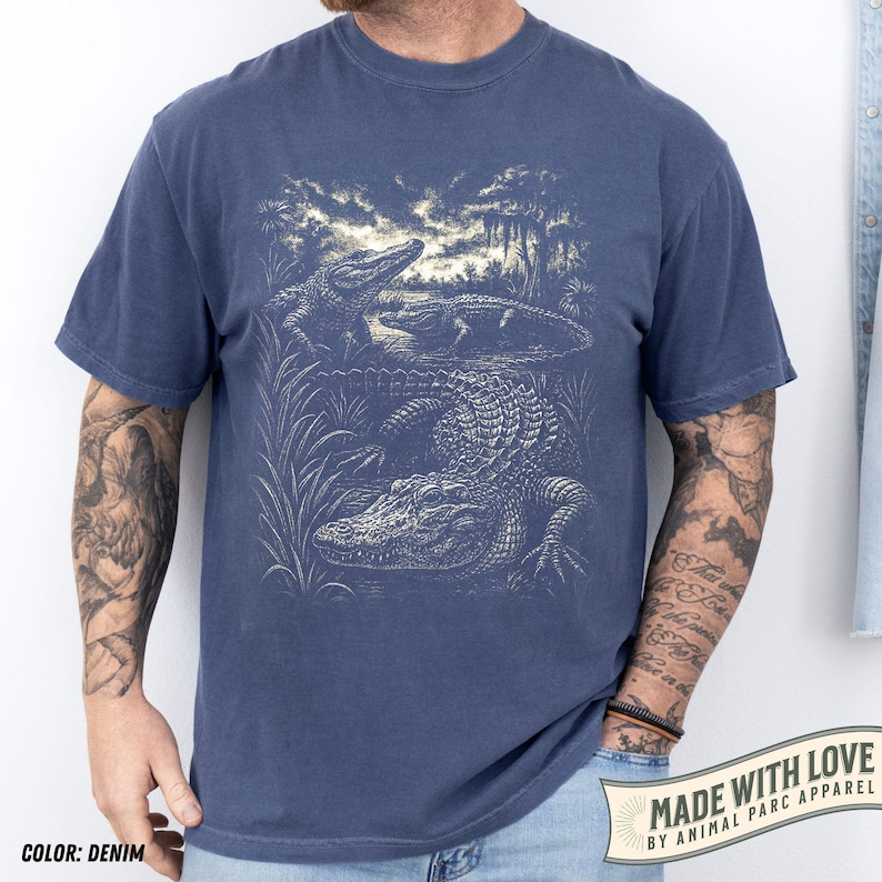 May include: Denim blue t-shirt featuring a detailed illustration of alligators in a swamp setting. The graphic is in a vintage style, with the text "MADE WITH LOVE BY ANIMAL PARC APPAREL" on a banner at the bottom right.