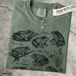 Könnte beinhalten: Moosgrünes T-Shirt mit detaillierten schwarzen Illustrationen verschiedener Froscharten, darunter Ochsenfrosch, Grünfrosch und Leopardfrosch. Das Shirt hat einen Rundhalsausschnitt und ein kleines Etikett mit der Aufschrift "COMFORT COLORS". Eine zarte Halskette mit Anhänger liegt auf dem Shirt.