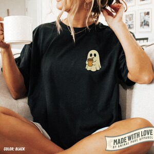 May include: Black t-shirt featuring a small, embroidered ghost holding a coffee cup. The ghost is beige with a friendly expression. The shirt has a relaxed fit and is being worn by a person. The words "MADE WITH LOVE" are visible.
