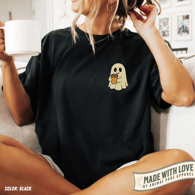 May include: Black t-shirt featuring a small, embroidered ghost holding a coffee cup. The ghost is beige with a friendly expression. The shirt has a relaxed fit and is being worn by a person. The words "MADE WITH LOVE" are visible.