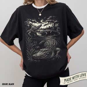 May include: Black t-shirt featuring a detailed illustration of alligators in a swamp setting. The design includes three alligators, tall grass, and trees. The text "MADE WITH LOVE BY ANIMAL PARC APPAREL" is printed on the bottom right corner. The shirt is black.