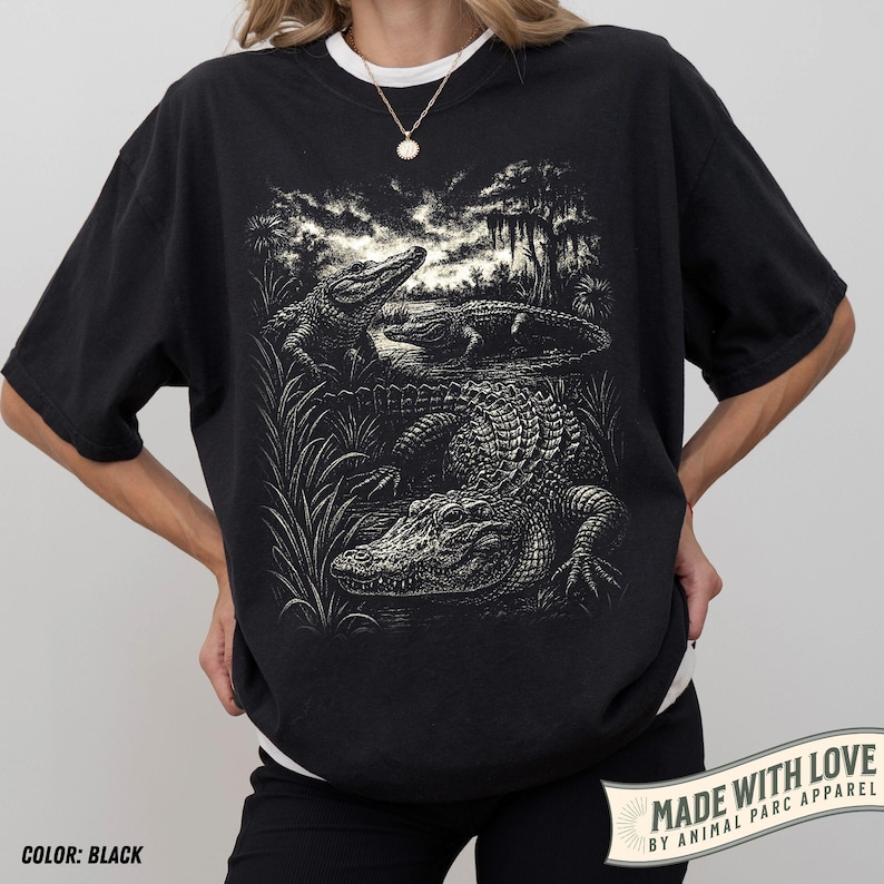 May include: Black t-shirt featuring a detailed illustration of alligators in a swamp setting. The design includes three alligators, tall grass, and trees. The text "MADE WITH LOVE BY ANIMAL PARC APPAREL" is printed on the bottom right corner. The shirt is black.