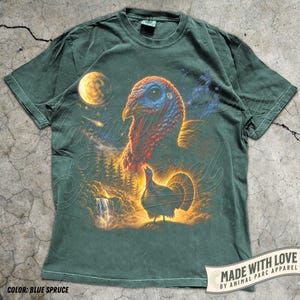 Puede incluir: Una camiseta de manga corta de color verde oscuro con un estampado gr&aacute;fico que muestra una cabeza de pavo grande en la parte superior y un pavo m&aacute;s peque&ntilde;o en un paisaje forestal con una cascada y una luna amarilla.