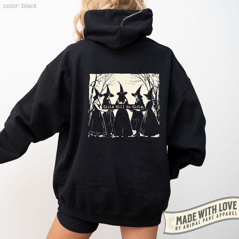 May include: Black hooded sweatshirt featuring a vintage-style graphic print on the back. The print depicts five witches in black robes and pointed hats, with the text 'Girls Will Be Girls.'  The sweatshirt is a pullover style with a large hood. The image is set against a plain white background.