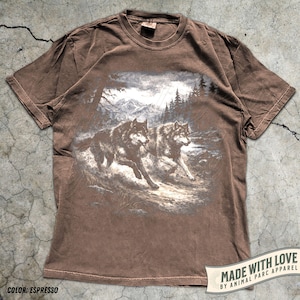 K&ouml;nnte beinhalten: Espressofarbenes T-Shirt mit einer detaillierten Grafik von zwei W&ouml;lfen, die durch eine Waldlandschaft laufen. Das Design umfasst Berge und B&auml;ume, mit dem Text "MADE WITH LOVE BY ANIMAL PARC APPAREL".