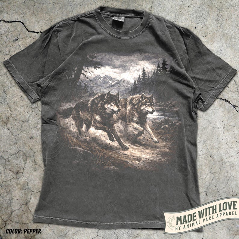 K&ouml;nnte beinhalten: Pfefferfarbenes T-Shirt mit einer detaillierten Grafik von zwei W&ouml;lfen, die durch eine Waldlandschaft mit Bergen im Hintergrund laufen. Das Shirt hat einen klassischen Rundhalsausschnitt und kurze &Auml;rmel. Der Text "MADE WITH LOVE BY ANIMAL PARC APPAREL" ist unten rechts aufgedruckt.