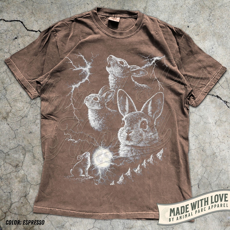 Pode incluir: Camiseta cor espresso com um gr&aacute;fico detalhado de coelhos, raios e lua cheia. O design inclui o texto "MADE WITH LOVE BY ANIMAL PARC APPAREL" e "COLOR: ESPRESSO".