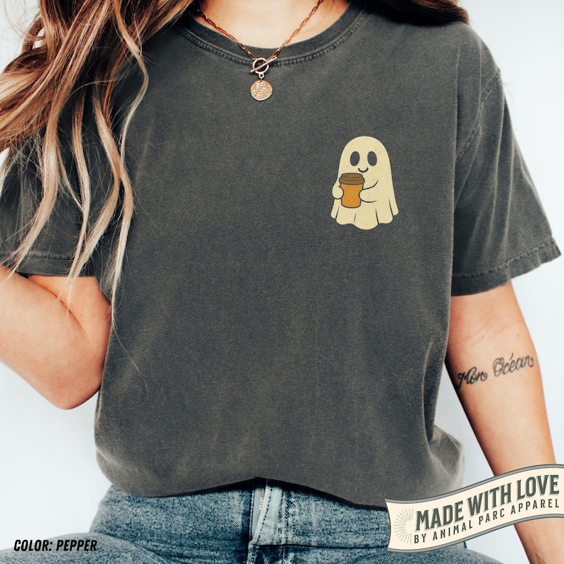 May include: Pepper-colored t-shirt featuring a friendly ghost holding a coffee cup. The ghost is light yellow with a smiling face, and the coffee cup is brown. The shirt has a relaxed fit and short sleeves. The text "MADE WITH LOVE BY ANIMAL PARC APPAREL" is at the bottom.