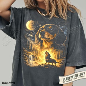 May include: Pepper-colored t-shirt featuring a detailed illustration of a dachshund dog under a starry night sky with a full moon. The design includes a waterfall and forest scene, with the text "MADE WITH LOVE BY ANIMAL PARC APPAREL".