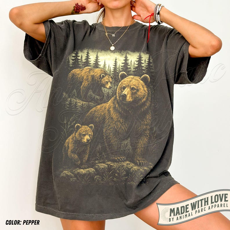 May include: Pepper-colored t-shirt featuring a detailed illustration of a bear family in a forest setting. The design includes a large bear with two cubs, set against a backdrop of trees and a sunset. The text "MADE WITH LOVE" is visible.