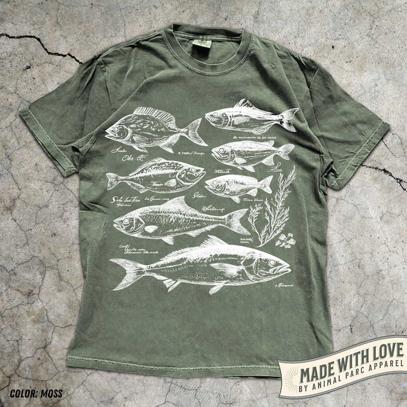 May include: Moss green t-shirt featuring a detailed white illustration of various fish species. The design includes scientific names and botanical elements. The shirt has a classic crew neck and short sleeves. The text "MADE WITH LOVE BY ANIMAL PARC APPAREL" is printed on a banner.