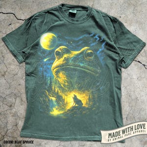May include: A dark green short-sleeve t-shirt featuring a graphic print of a large frog, a bright yellow moon, a starry night sky, and a silhouette of a wolf howling near a waterfall. The shirt has a crew neckline and a standard fit.