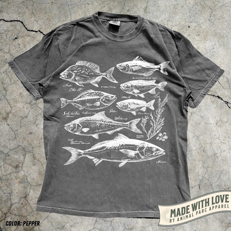 May include: Pepper-colored t-shirt featuring a detailed white illustration of various fish species. The design includes scientific names and botanical elements. The shirt has a classic crew neck and short sleeves. The text "MADE WITH LOVE BY ANIMAL PARC APPAREL" is printed on the bottom right.