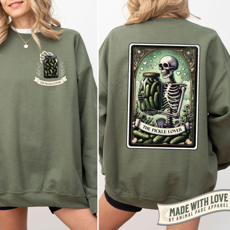 May include: Olive green sweatshirt with a pickle-themed design. The front features a jar of pickles with the text 'THE PICKLE LOVER'. The back displays a tarot card design with a skeleton holding a pickle jar, surrounded by pickles and the same text. The design is framed with a decorative border.