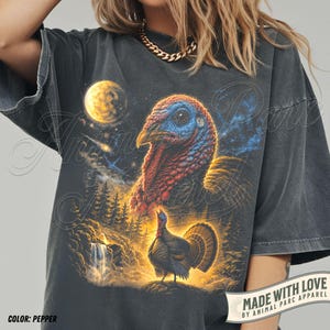 Puede incluir: Una camiseta gris carb&oacute;n con un estampado gr&aacute;fico de la cabeza de un pavo y un pavo de cuerpo entero en un paisaje forestal con una cascada y una luna brillante en el cielo nocturno. La camiseta tiene un aspecto desgastado y mangas cortas.