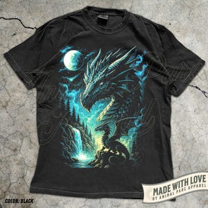 May include: Black t-shirt featuring a detailed dragon illustration. The design includes a dragon's head, a smaller dragon, a waterfall, and a full moon in a night sky. The shirt has the text "MADE WITH LOVE BY ANIMAL PARC APPAREL" and "COLOR: BLACK".