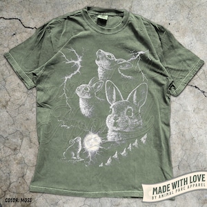 Pode incluir: T-shirt verde musgo com um design gr&aacute;fico detalhado de coelhos, uma lua cheia e raios. O design inclui o texto "Animal Apparel" e "Made with Love by Animal Parc Apparel".