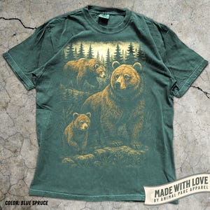 May include: Blue spruce t-shirt featuring a detailed illustration of a bear family in a forest setting. The design includes a large bear and two cubs, with a backdrop of pine trees. The shirt has a vintage look and feel.