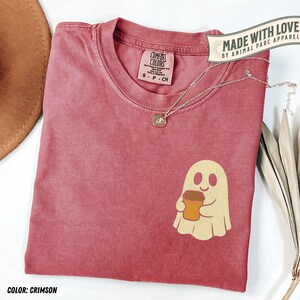 May include: Crimson Comfort Colors t-shirt with a ghost holding a coffee cup graphic. The shirt has a crew neck and short sleeves. A silver necklace with a small pendant is on the shirt. The text "MADE WITH LOVE BY ANIMAL PARC APPAREL" is on the top right.