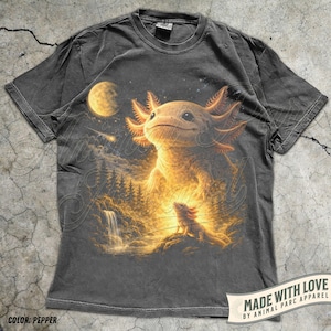 May include: Pepper-coloured t-shirt with a detailed illustration of an axolotl in a fantasy landscape with a waterfall, forest, and moon. The design includes the text "MADE WITH LOVE BY ANIMAL PARC APPAREL" on a banner. The shirt's colour is described as "PEPPER".