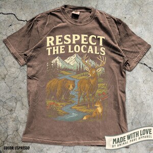 May include: Espresso-colored t-shirt with the text "Respect the Locals" above a nature scene. The graphic features a bear, deer, and fox near a river and mountains. Made with love by Animal Parc Apparel.