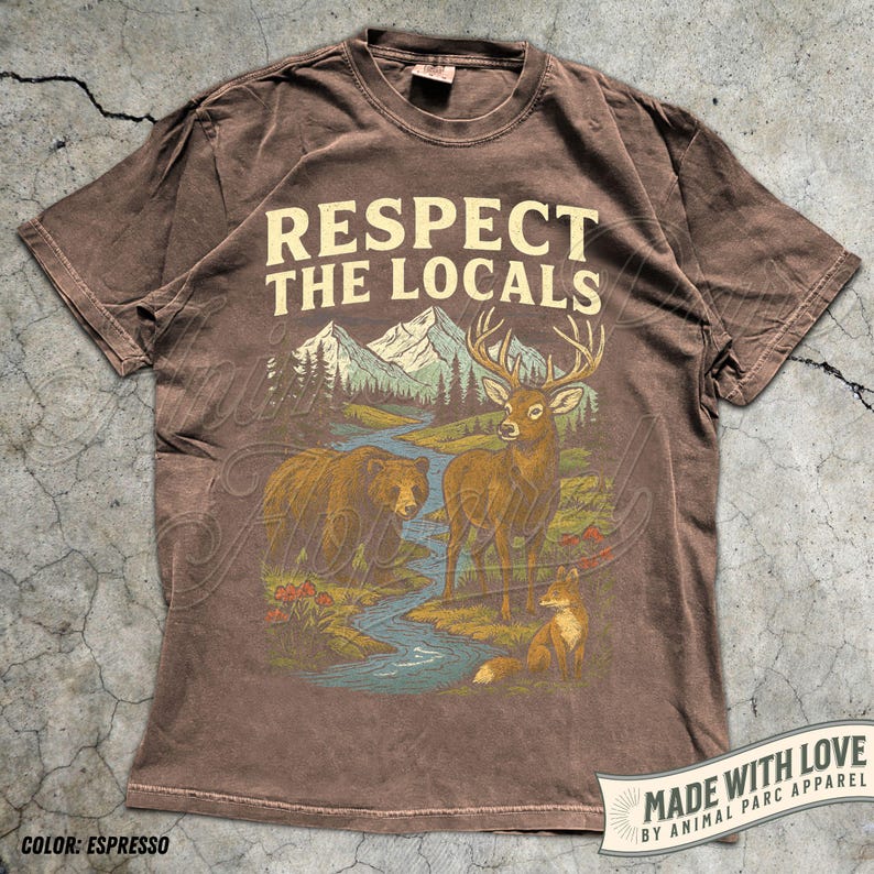 May include: Espresso-colored t-shirt with the text "Respect the Locals" above a nature scene. The graphic features a bear, deer, and fox near a river and mountains. Made with love by Animal Parc Apparel.