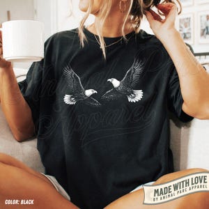 Może przedstawiać: Czarny t-shirt z białą grafiką przedstawiającą dwa orły w locie. Koszulka ma napis "Animal Parc Apparel" i "Made with Love by Animal Parc Apparel".