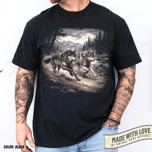 K&ouml;nnte beinhalten: Schwarzes T-Shirt mit einer detaillierten Grafik von zwei W&ouml;lfen, die durch eine Waldlandschaft mit Bergen im Hintergrund laufen. Das T-Shirt enth&auml;lt den Text "MADE WITH LOVE BY ANIMAL PARC APPAREL".