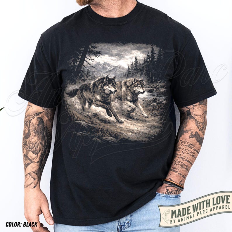 K&ouml;nnte beinhalten: Schwarzes T-Shirt mit einer detaillierten Grafik von zwei W&ouml;lfen, die durch eine Waldlandschaft mit Bergen im Hintergrund laufen. Das T-Shirt enth&auml;lt den Text "MADE WITH LOVE BY ANIMAL PARC APPAREL".