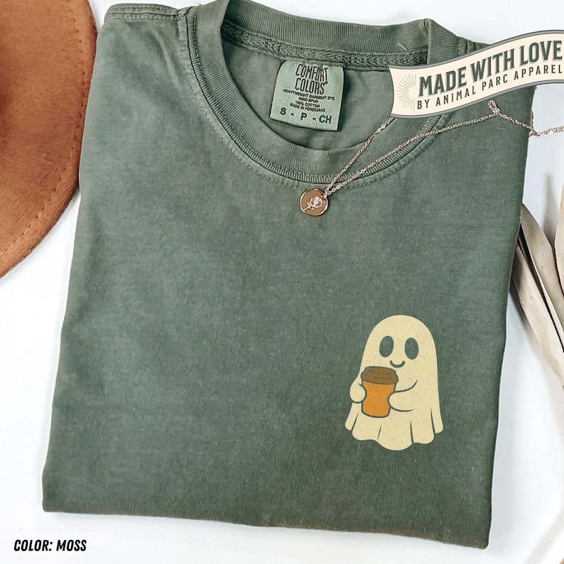 May include: A moss green Comfort Colors t-shirt with a ghost graphic holding a coffee cup. The shirt has a small tag and a necklace. The text "MADE WITH LOVE BY ANIMAL PARC APPAREL" is visible.