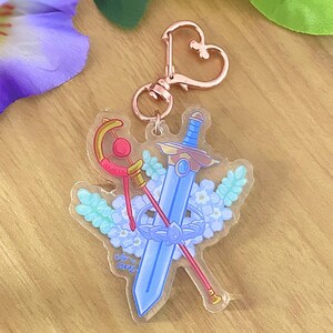 May include: Clear acrylic keychain with a blue sword, red and gold staff, and floral design. It has a rose gold heart-shaped clasp. The design includes the text "Shira Gartz".