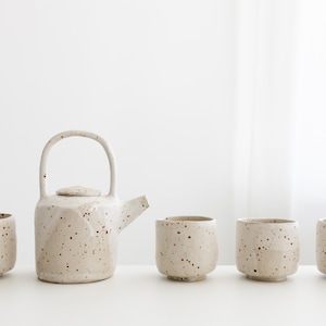 Op de afbeelding: Een witte theepot van keramiek en vier bijpassende kopjes met een gespikkeld glazuur. De theepot heeft een rond lichaam en een tuit aan de zijkant. De kopjes zijn kort en breed.