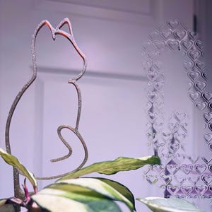 May include: A decorative cat-shaped plant stake with glittery accents, placed in a pot with a houseplant. The stake is a silhouette of a cat. The plant has green and white leaves. A heart-shaped pattern is visible in the background.