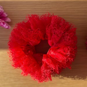 Könnte beinhalten: Ein leuchtend rotes Scrunchie aus zarter Spitzenstoff, mittig auf einer hellen Holzoberfläche platziert. Zwei rosa Blüten, eine Gerbera und eine dekorative Blüte, befinden sich auf beiden Seiten des Scrunchies. Die Textur des Scrunchies kontrastiert mit dem glatten Holz.