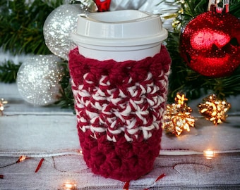 May include: A red and white crocheted coffee cup cosy with a white paper cup inside. The cosy is surrounded by silver and gold baubles and a green pine branch.