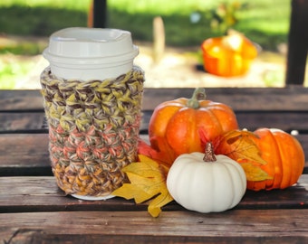 May include: A crocheted coffee cup cozy in shades of brown, orange, and yellow, with fall-themed pumpkins and leaves.