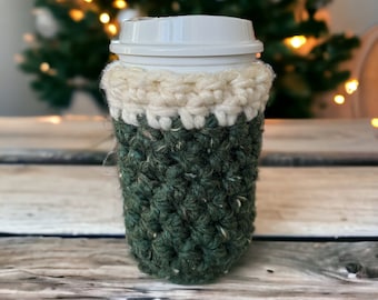 May include: A green and white crocheted coffee cup cozy. The cozy is made with a textured yarn and has a white band around the top.