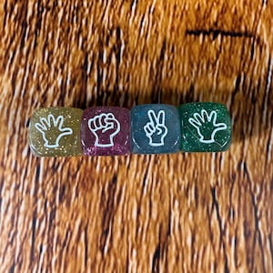 May include: Four colorful resin dice with white hand gestures: open hand, fist, peace sign, and open hand. The dice are gold, magenta, teal, and green, with glitter accents. The background is a brown, textured surface.
