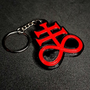 Pode incluir: Um chaveiro preto e vermelho com um símbolo que parece uma cruz e um sinal de infinito. O símbolo é impresso em 3D e tem um contorno vermelho.