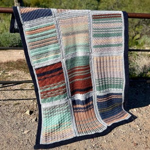 May include: A handmade patchwork blanket with a navy blue border. The blanket features a variety of textured squares in shades of blue, green, brown, and peach. The blanket is draped over a metal railing with a desert landscape in the background.