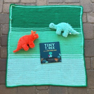 May include: A green, blue, and white crocheted baby blanket with a book titled "Tiny T. Rex and the Very Dark Dark" and two plush dinosaur toys.