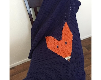 May include: A navy blue crocheted blanket with an orange fox face design. The blanket is draped over a wooden chair.