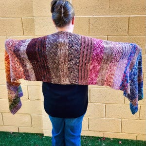 Pode incluir: Uma echarpe de crochê colorida com uma variedade de cores, incluindo roxo, rosa, azul e marrom. A echarpe está drapeada sobre os ombros de uma pessoa que está vestindo uma camisa preta e calças jeans azuis.