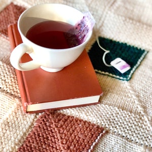May include: A white teacup with a tea bag resting on the rim, filled with a dark red liquid. The teacup is sitting on top of a brown book, which is resting on a white knitted blanket with brown and green squares.
