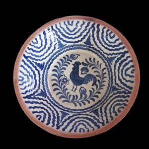 Op de afbeelding: Een ronde keramische schaal met een blauw en wit hanenontwerp. De schaal heeft een bruine rand en een centrale haan omringd door bloemmotieven. De buitenrand heeft een herhalend blauw golfpatroon.
