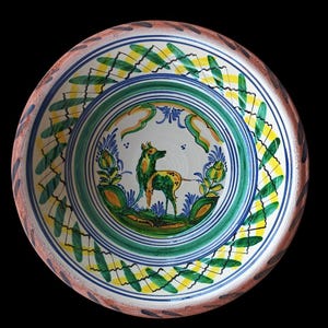 May include: A hand-painted ceramic plate with a deer motif. The plate features a white center with a green deer, surrounded by green, blue, and yellow floral designs. The rim has a brown and white pattern, and the plate is set against a black background.