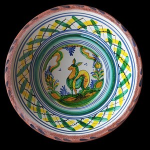May include: A hand-painted ceramic bowl with a deer design. The bowl features a brown rim, blue and yellow geometric patterns, and a central image of a deer in green and yellow. The background is black.